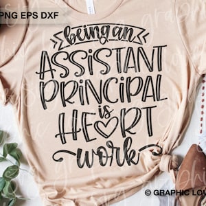 May include: A beige t-shirt with the text "Being an Assistant Principal is Heart Work" in black lettering. The text is in a stylized font with a heart symbol in the word "Heart".