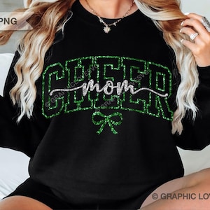 Könnte beinhalten: Ein schwarzes Langarm-Sweatshirt mit dem Wort CHEER in großen, grün glitzernden Blockbuchstaben und dem darüber liegenden Wort mom in silberner Schreibschrift, ergänzt durch eine kleine, grün glitzernde Schleifengrafik unter dem Text.