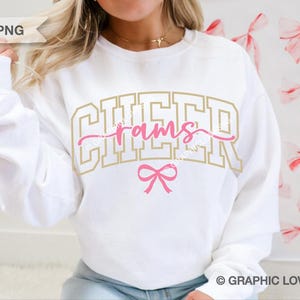 May include: White sweatshirt with the word "CHEER" in gold block letters and "rams" in pink script. A pink bow is centered below the text. The sweatshirt is a casual style, suitable for sports fans.