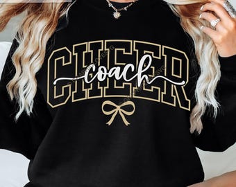 Puede incluir: Sudadera negra con la palabra "CHEER" en letras doradas y la palabra "coach" en escritura blanca. Un lazo dorado debajo. Diseño temático de animadoras o entrenadores.