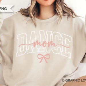 May include: A beige sweatshirt featuring the text "DANCE MOM" in white block letters and pink script, with a pink bow beneath. The sweatshirt is a relaxed fit, suitable for dance mums.