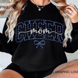 Peut inclure: Sweat-shirt noir avec inscription "Cheer Mom" en paillettes bleues et un nœud en paillettes argentées en dessous.