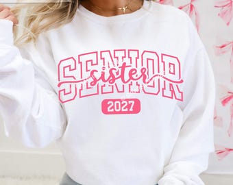 Peut inclure: Un sweat-shirt blanc à manches longues avec une inscription rose de style universitaire SENIOR, le mot sister en écriture cursive superposé au centre et le nombre 2027 imprimé dans un rectangle rose situé sous le texte principal.