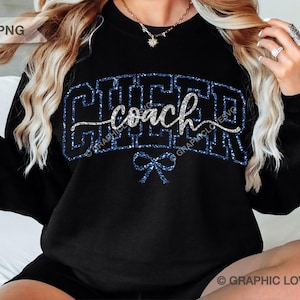 May include: Black sweatshirt with the word "CHEER" in blue glitter letters and the word "coach" in silver script. A blue glitter bow is below the word "coach". The sweatshirt is a crew neck style.