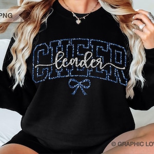 Op de afbeelding: Zwart sweatshirt met het woord "CHEER" in een blauw glitterlettertype, met "leader" in een cursief lettertype en een blauwe strik. De sweatshirt heeft een ronde hals.