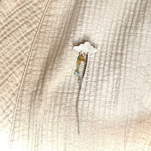 Op de afbeelding: Een witte, wolk-vormige broche met een bungelende bedel. De bedel heeft een klein figuur met gouden en turquoise accenten, hangend aan een zilveren ketting. De broche is vastgemaakt aan een crèmekleurige, gewatteerde stof.