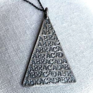 May include: A silver triangular pendant featuring the word "ABRACADABRA" repeatedly engraved. The pendant is suspended from a fine, dark chain. The design is simple but striking, suitable for everyday wear. The pendant is approximately 3 cm in height.