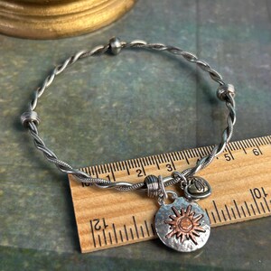 Pu&ograve; includere: Un braccialetto rigido in filo ritorto argento e rame con un ciondolo a forma di sole e un ciondolo a forma di cuore. Il braccialetto ha un diametro di circa 15 cm.