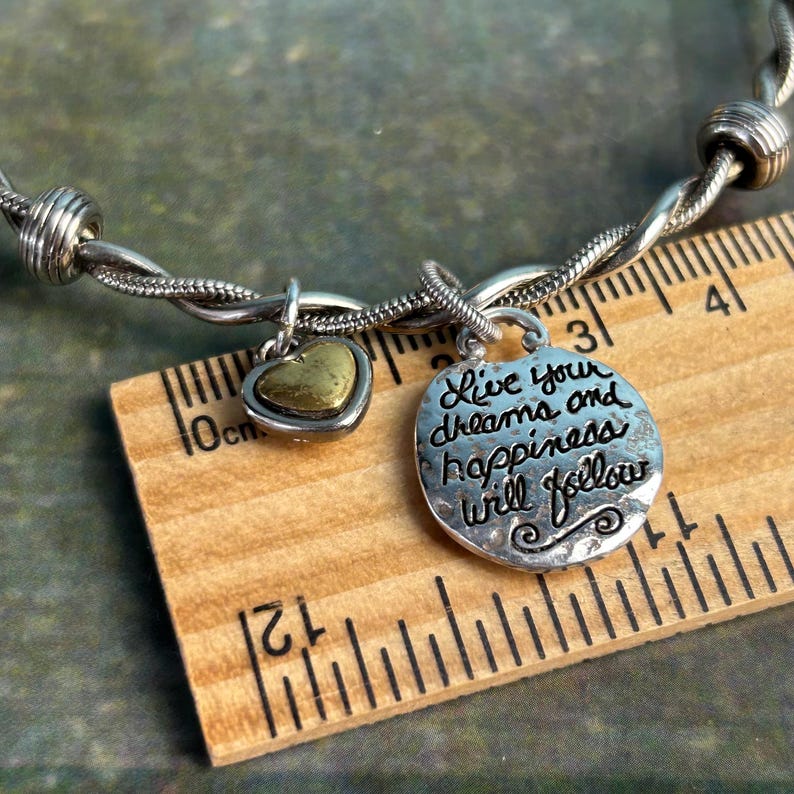 Pu&ograve; includere: Braccialetto d'argento con un ciondolo a forma di cuore e un ciondolo che dice "Live your dreams and happiness will follow". Il braccialetto viene misurato contro un righello di legno.