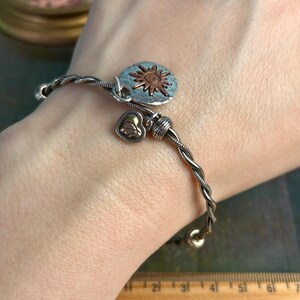 Pu&ograve; includere: Un braccialetto rigido intrecciato in argento e rame con un ciondolo a forma di sole e un ciondolo a forma di cuore.