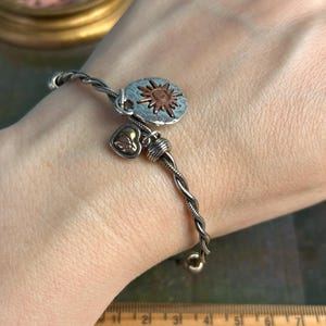 Pu&ograve; includere: Un braccialetto in argento e rame con un ciondolo a forma di sole e un ciondolo a forma di cuore. Il braccialetto &egrave; intrecciato e ha un design delicato.