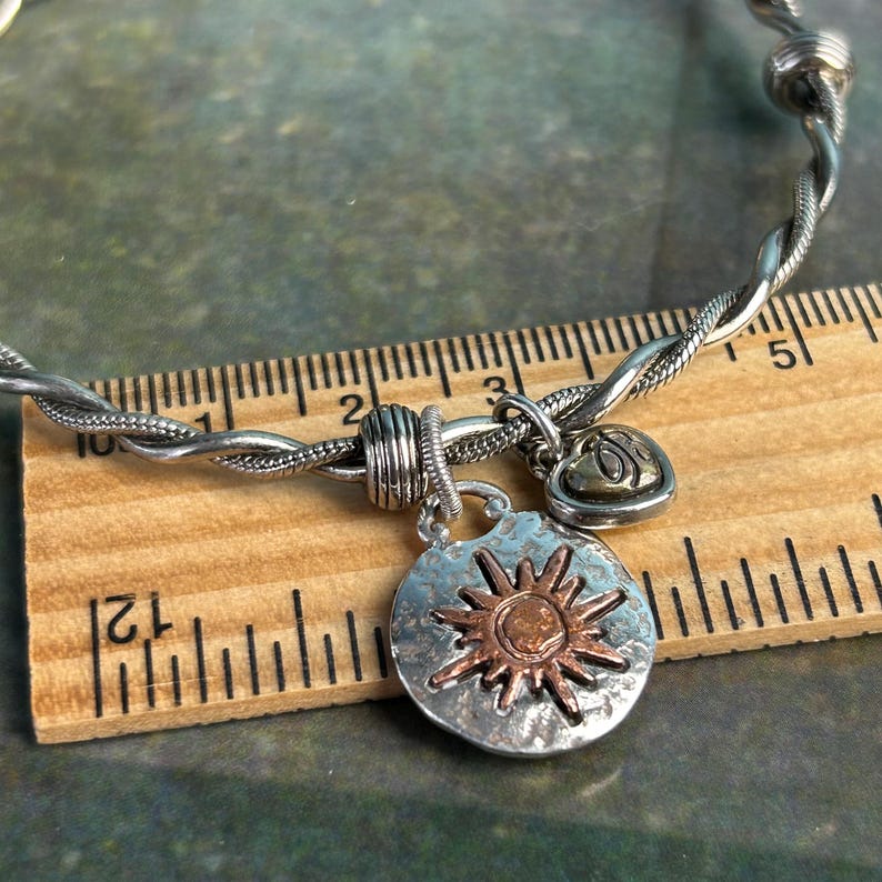 Pu&ograve; includere: Un braccialetto in argento e rame con un ciondolo a forma di sole e un ciondolo a forma di cuore. Il braccialetto &egrave; intrecciato e misura circa 18 cm di lunghezza.