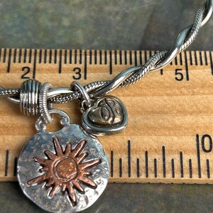 Pu&ograve; includere: Un braccialetto d'argento con un design intrecciato e tre ciondoli. I ciondoli sono un cuore con un design a spirale, un sole con un centro in rame e un disco d'argento con una superficie testurizzata.