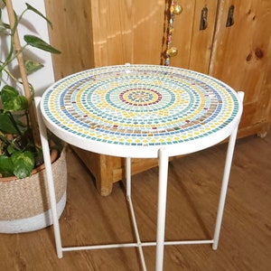 Puede incluir: Una mesa auxiliar de metal blanco con una tapa redonda de mosaico. El mosaico está hecho de pequeñas baldosas de colores en tonos azules, verdes, amarillos y rojos. La mesa tiene un diseño simple y moderno.