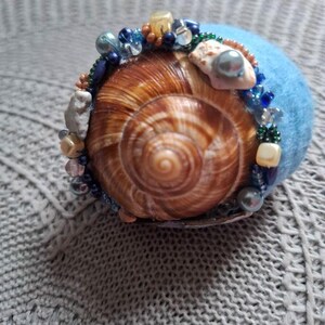 Könnte beinhalten: Dekoratives Schneckenhaus, verziert mit blauen, klaren und perlmuttfarbenen Perlen, liegt auf einem blauen Filzsockel. Das Gehäuse hat ein warmes Braun mit Spiralmustern. Zusätzliche kleine Muscheln und Steine sind integriert.