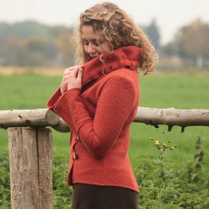 Könnte beinhalten: Eine Frau trägt eine rostfarbene, langärmelige Jacke mit hohem Kragen. Die Jacke hat einen Knopfverschluss und ist aus einem weichen, strukturierten Stoff gefertigt. Die Frau steht auf einem Feld mit einem Holzzaun hinter ihr.