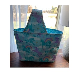 May include: A light blue tote bag with a handle, featuring a pattern of purple and blue unicorns with rainbows on a white background.