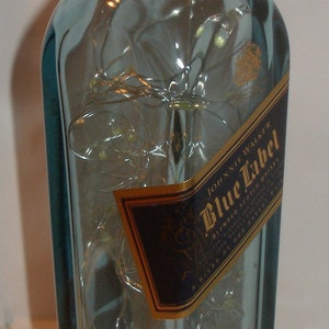 K&ouml;nnte beinhalten: Eine durchsichtige Glasflasche mit einem goldenen Verschluss und einem blauen Etikett mit der Aufschrift "Johnnie Walker Blue Label Blended Scotch Whisky" und einem goldenen Siegel mit einem laufenden Mann.