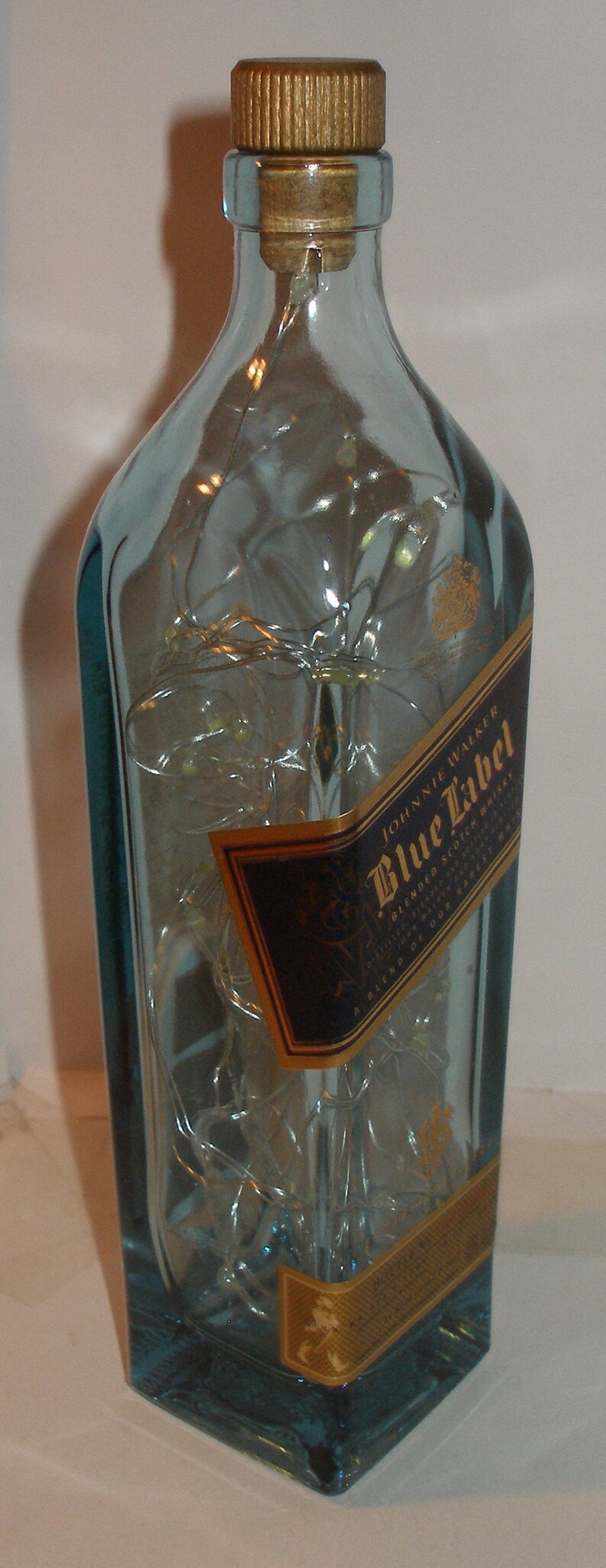 K&ouml;nnte beinhalten: Eine durchsichtige Glasflasche mit einem goldenen Verschluss und einem blauen Etikett mit der Aufschrift "Johnnie Walker Blue Label Blended Scotch Whisky" und einem goldenen Siegel mit einem laufenden Mann.