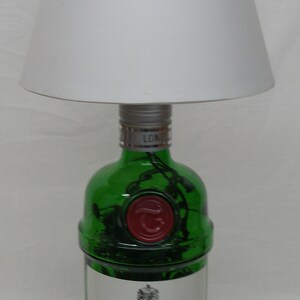 May include: A green glass bottle with a white label that reads "Tanqueray Imported London Dry Gin". The bottle is topped with a silver cap and has a red wax seal. The bottle is sitting on a white surface under a white lampshade.