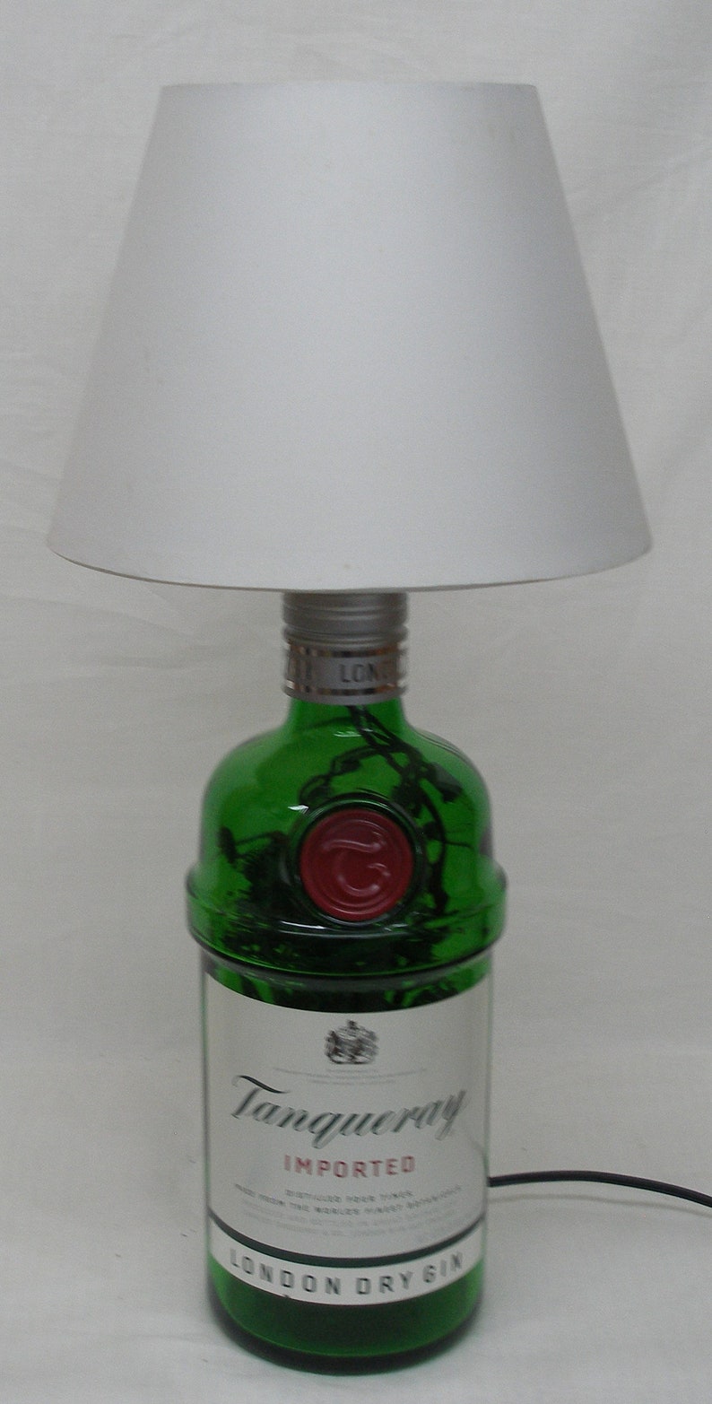 May include: A green glass bottle with a white label that reads "Tanqueray Imported London Dry Gin". The bottle is topped with a silver cap and has a red wax seal. The bottle is sitting on a white surface under a white lampshade.