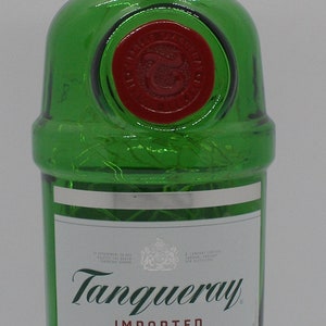 Puede incluir: Una botella de vidrio verde de Tanqueray London Dry Gin. La botella tiene una tapa plateada con las palabras "London Dry Gin" impresas en ella. La etiqueta es blanca con texto negro y un escudo plateado. El texto en la etiqueta dice "Tanqueray Importado, Destilado Cuatro Veces, Hecho con los Mejores Bot&aacute;nicos del Mundo, Gin Seco de Londres, Producido y Embotellado en Gran Breta&ntilde;a para Charles Tanqueray & Co., 47.3% Alc. vol. 194.6 Prueba, 375 ml."