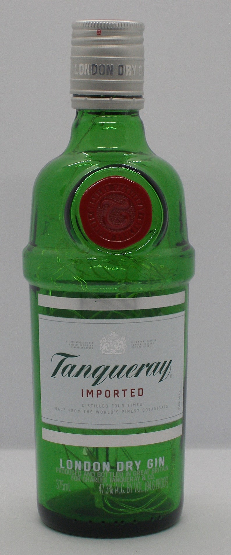 Puede incluir: Una botella de vidrio verde de Tanqueray London Dry Gin. La botella tiene una tapa plateada con las palabras "London Dry Gin" impresas en ella. La etiqueta es blanca con texto negro y un escudo plateado. El texto en la etiqueta dice "Tanqueray Importado, Destilado Cuatro Veces, Hecho con los Mejores Bot&aacute;nicos del Mundo, Gin Seco de Londres, Producido y Embotellado en Gran Breta&ntilde;a para Charles Tanqueray & Co., 47.3% Alc. vol. 194.6 Prueba, 375 ml."