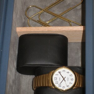 May include: A gold watch with a white face and black hands. The watch is in a black leather watch box. There are two gold paper clips in the top drawer.