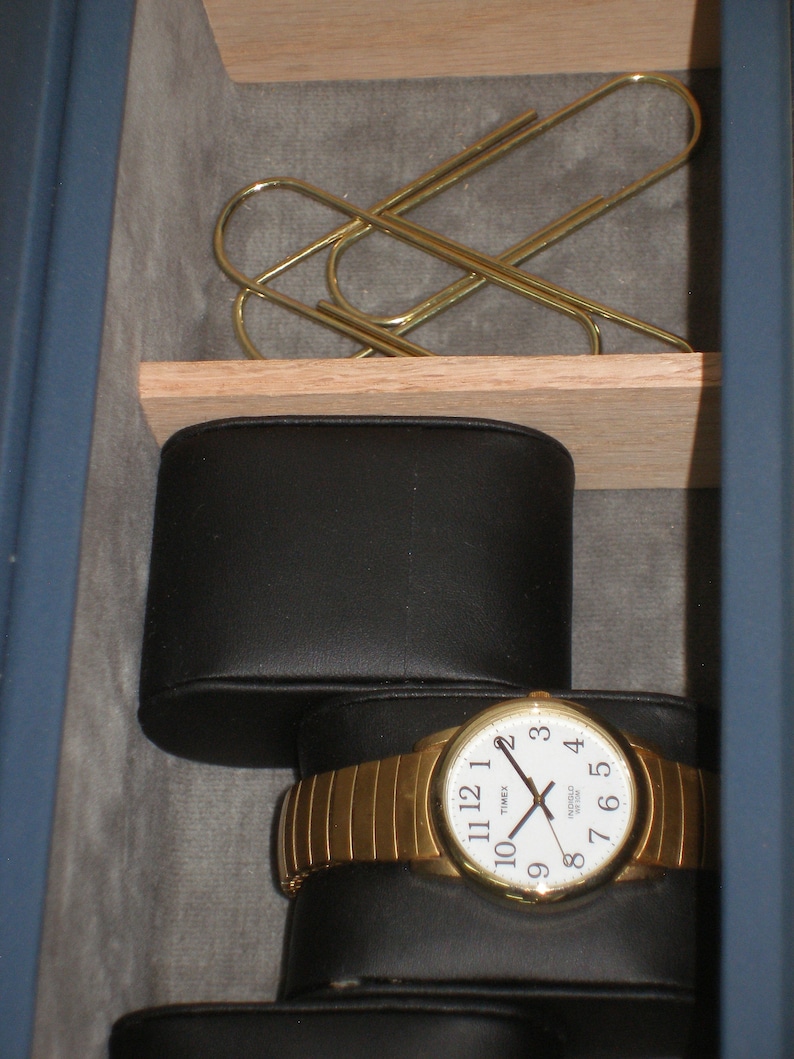 May include: A gold watch with a white face and black hands. The watch is in a black leather watch box. There are two gold paper clips in the top drawer.