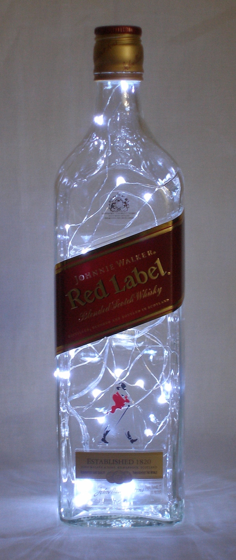 May include: A clear glass bottle with a red label that says "Johnnie Walker Red Label Blended Scotch Whisky Established 1820" with white string lights wrapped around it.