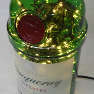 May include: A green glass bottle with a red wax seal and a white label that reads "Tanqueray Imported London Dry Gin". The bottle is filled with string lights.