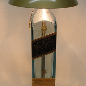 Pu&ograve; includere: Una lampada da tavolo unica realizzata con una bottiglia di whisky Johnnie Walker Blue Label riciclata. La lampada presenta un paralume a cupola verde metallizzato, una base in metallo spazzolato e una catena dorata. L'etichetta della bottiglia &egrave; visibile.