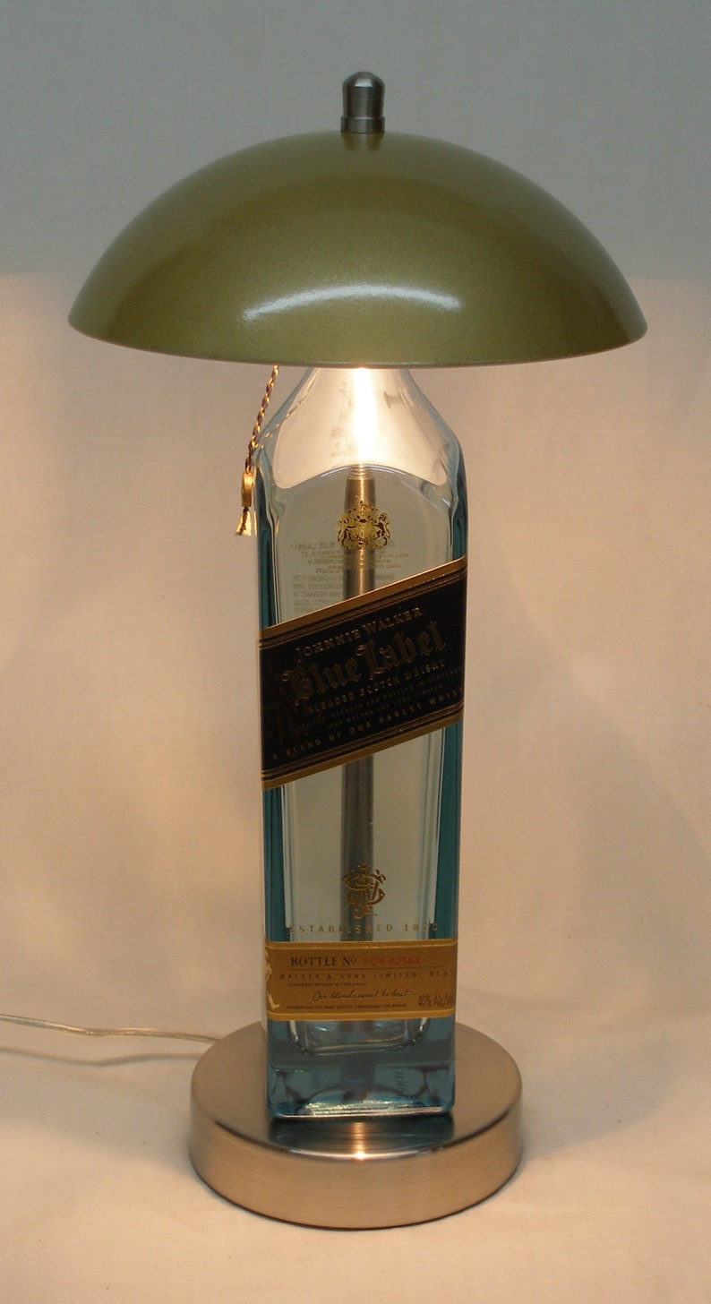 Pu&ograve; includere: Una lampada da tavolo unica realizzata con una bottiglia di whisky Johnnie Walker Blue Label riciclata. La lampada presenta un paralume a cupola verde metallizzato, una base in metallo spazzolato e una catena dorata. L'etichetta della bottiglia &egrave; visibile.