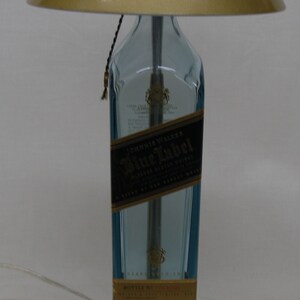 Pu&ograve; includere: Una lampada da tavolo unica realizzata con una bottiglia riciclata di Johnnie Walker Blue Label. La lampada presenta una base in vetro blu chiaro, un paralume dorato e una base argentata. Un elemento decorativo creativo per la casa.