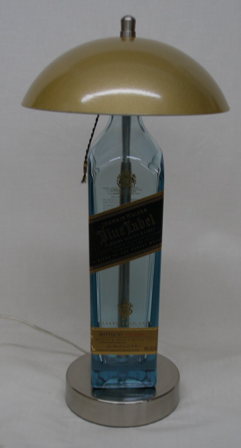 Pu&ograve; includere: Una lampada da tavolo unica realizzata con una bottiglia riciclata di Johnnie Walker Blue Label. La lampada presenta una base in vetro blu chiaro, un paralume dorato e una base argentata. Un elemento decorativo creativo per la casa.