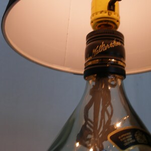 May include: A repurposed glass bottle lamp with a black lampshade and a gold light switch. The bottle has a label that reads "Johnnie Walker" and is clear with a textured surface.