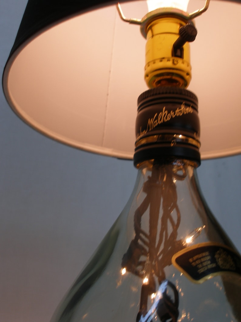May include: A repurposed glass bottle lamp with a black lampshade and a gold light switch. The bottle has a label that reads "Johnnie Walker" and is clear with a textured surface.