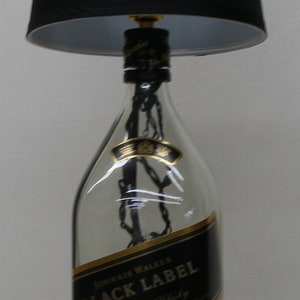 May include: A black lampshade sits atop a clear glass bottle with a gold label that reads "Johnnie Walker Black Label Blended Scotch Whisky". The bottle is filled with black string lights.