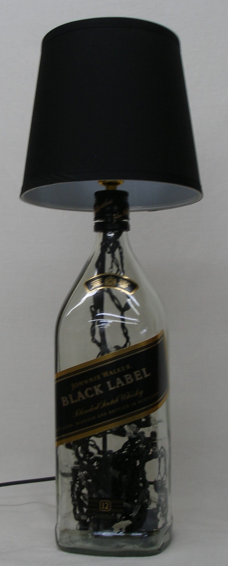 May include: A black lampshade sits atop a clear glass bottle with a gold label that reads "Johnnie Walker Black Label Blended Scotch Whisky". The bottle is filled with black string lights.