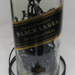 May include: A clear glass bottle with a black label that reads "JOHNNIE WALKER BLACK LABEL Blended Scotch Whisky DISTILLED, BLENDED AND BOTTLED IN SCOTLAND". The bottle is filled with black string lights and has a gold label with the text "12" on it.