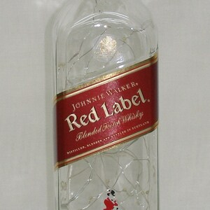 May include: A clear glass bottle of Johnnie Walker Red Label Blended Scotch Whisky. The label is red with gold lettering and features the Johnnie Walker logo of a man in a walking pose. The bottle is decorated with a string of white lights.
