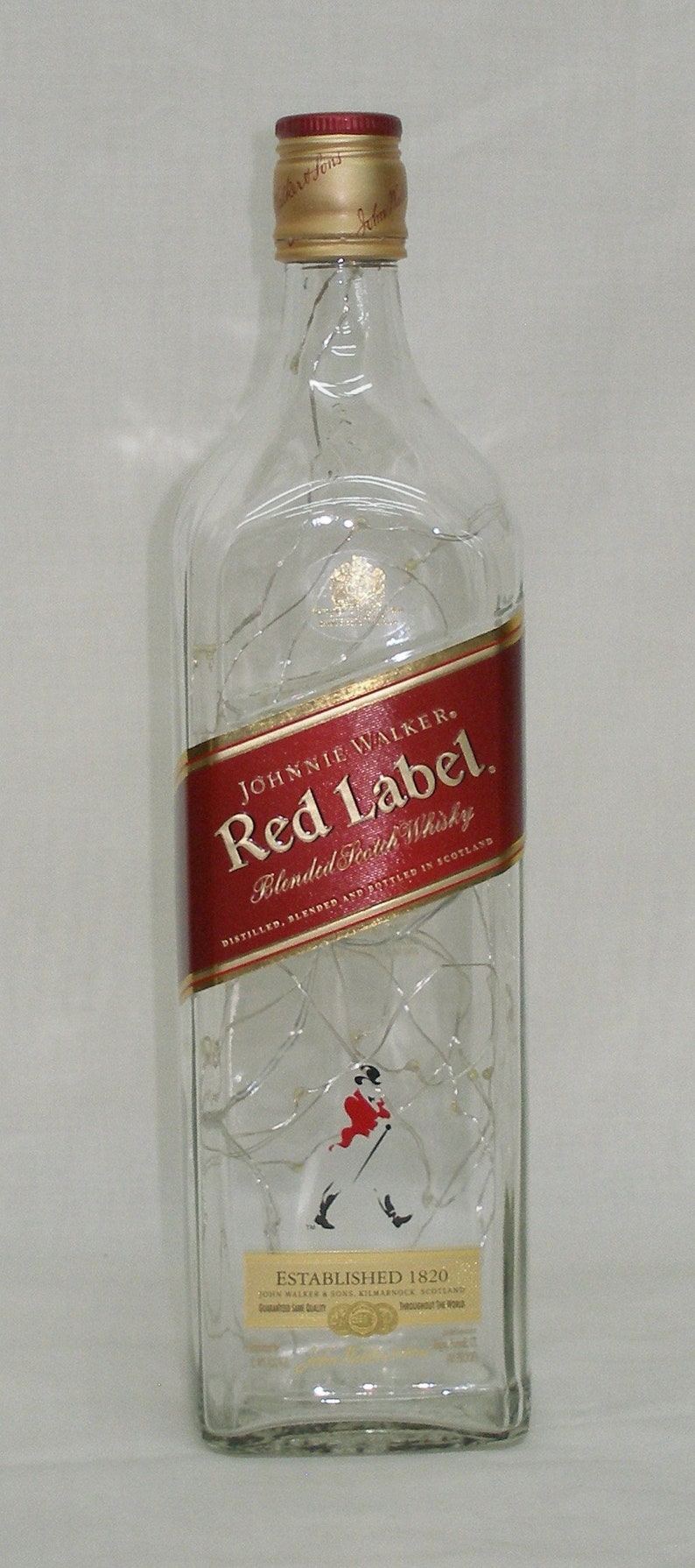 May include: A clear glass bottle of Johnnie Walker Red Label Blended Scotch Whisky. The label is red with gold lettering and features the Johnnie Walker logo of a man in a walking pose. The bottle is decorated with a string of white lights.