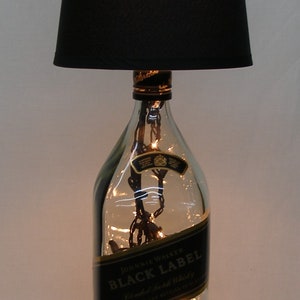 May include: A black lampshade with a clear glass bottle base. The bottle is filled with fairy lights and has a label that reads "Johnnie Walker Black Label Blended Scotch Whisky".