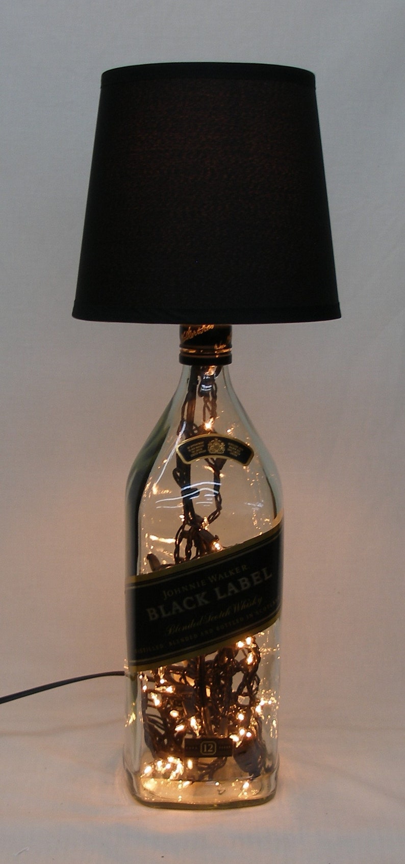 May include: A black lampshade with a clear glass bottle base. The bottle is filled with fairy lights and has a label that reads "Johnnie Walker Black Label Blended Scotch Whisky".