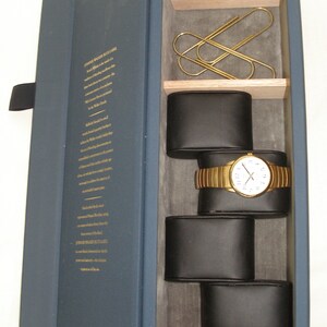 May include: A navy blue box with a gold inscription on the front. The box is open, revealing a watch with a gold band and white face, resting on a black cushion. The watch is surrounded by other black cushions, and there are three gold paper clips in the top compartment.