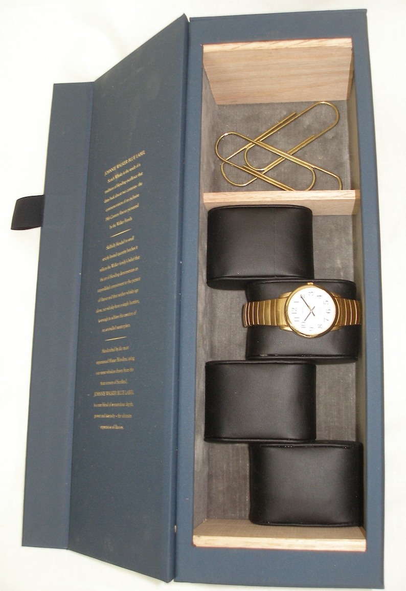May include: A navy blue box with a gold inscription on the front. The box is open, revealing a watch with a gold band and white face, resting on a black cushion. The watch is surrounded by other black cushions, and there are three gold paper clips in the top compartment.