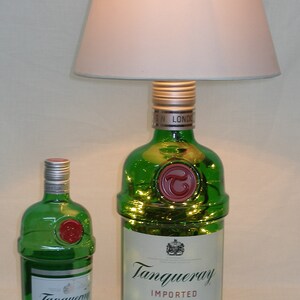 May include: Two green glass bottles of Tanqueray London Dry Gin. The larger bottle is a lamp with a white lampshade. The smaller bottle is a standard size bottle.