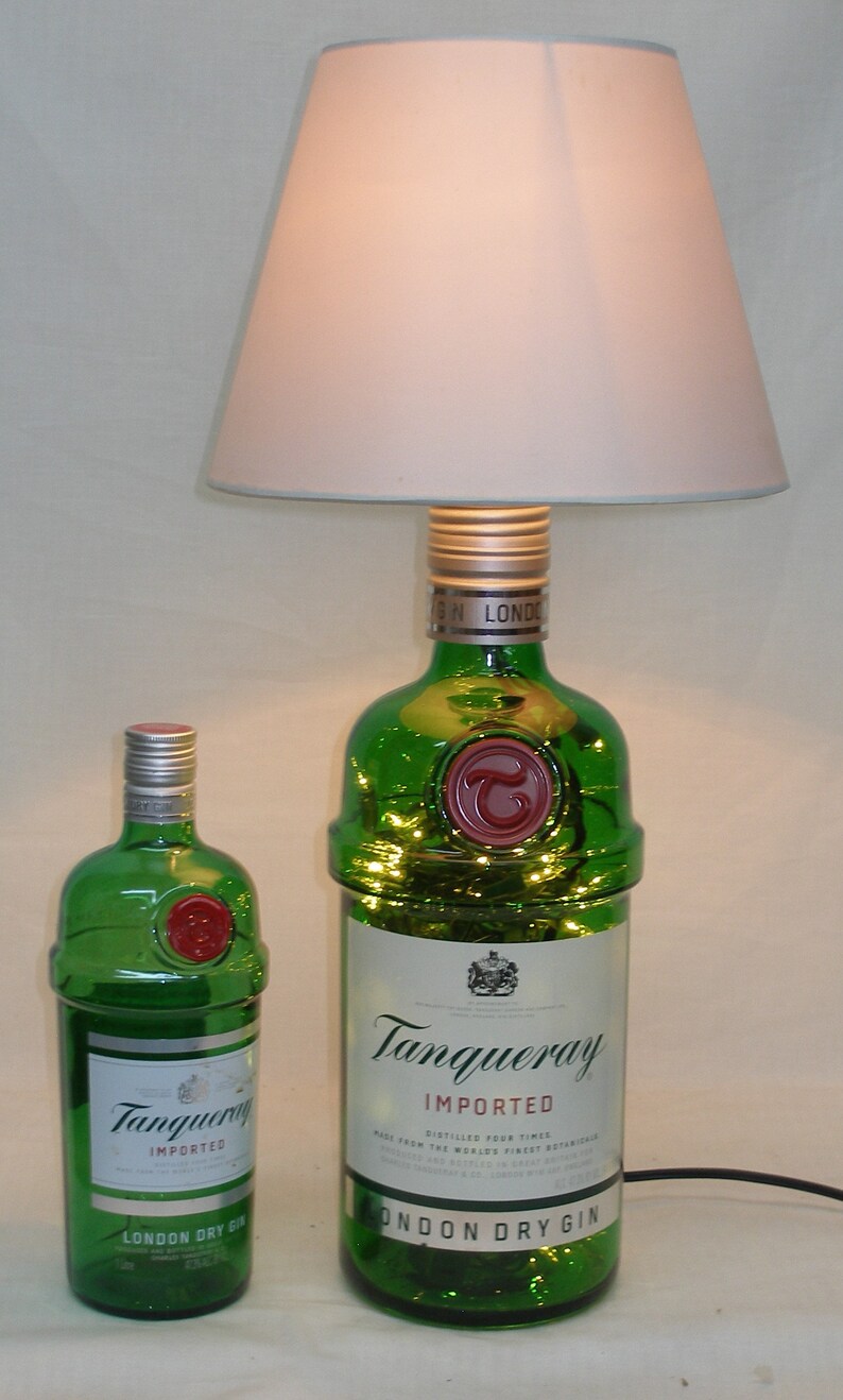May include: Two green glass bottles of Tanqueray London Dry Gin. The larger bottle is a lamp with a white lampshade. The smaller bottle is a standard size bottle.