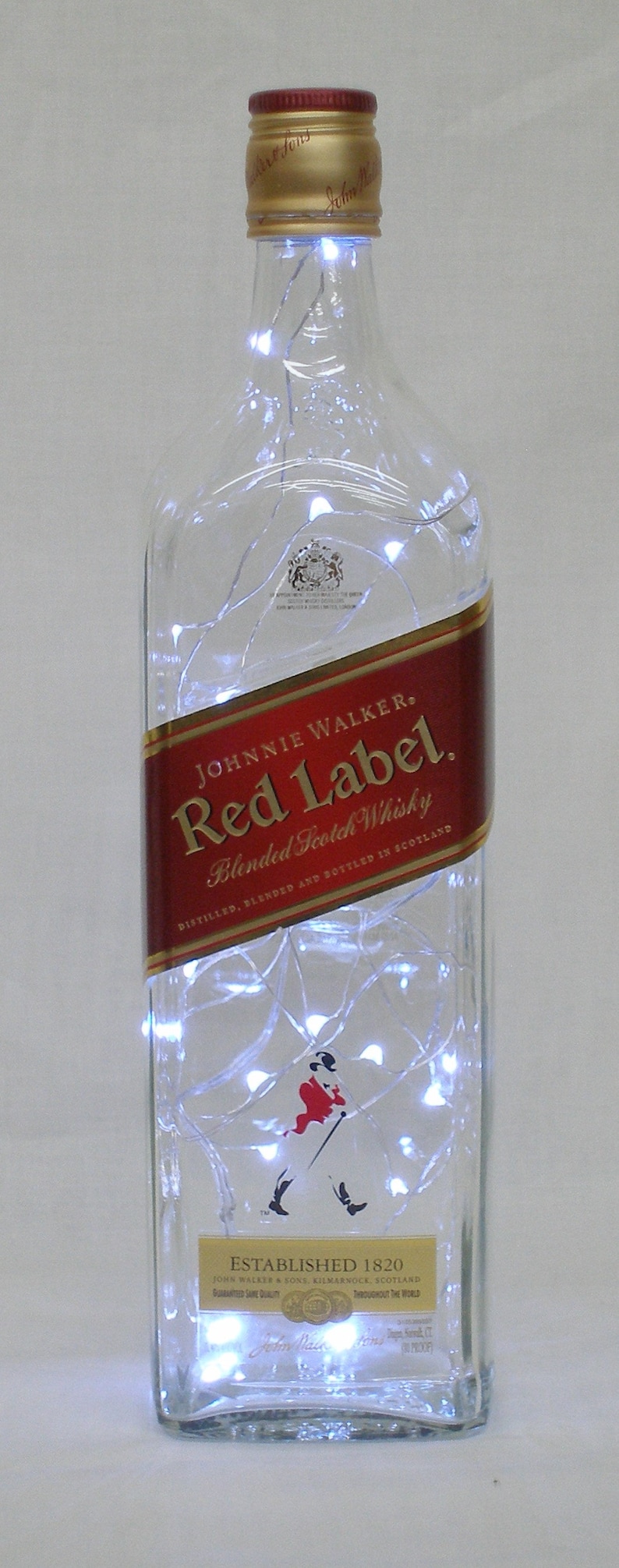 May include: A clear glass bottle with a red label that says "JOHNNIE WALKER. Red Label. Blended Scotch Whisky" and a gold label that says "ESTABLISHED 1820". The bottle is filled with white string lights.