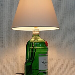 May include: A white lampshade with a green glass bottle base. The bottle is labeled "Tanqueray" and has a red wax seal. The lamp is turned on and illuminating the room.